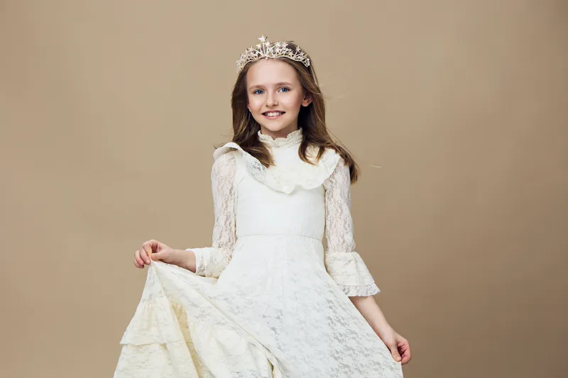 Young girl in a white princess dress with lace details and a crown, smiling cheerfully against a soft beige background, ideal for concepts of childhood and celebrations