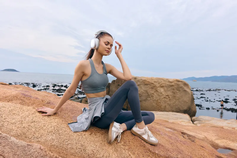 Young woman in athletic wear relaxing on rocky beach, enjoying music with headphones, peaceful atmosphere, serene emotions, outdoor lifestyle concept
