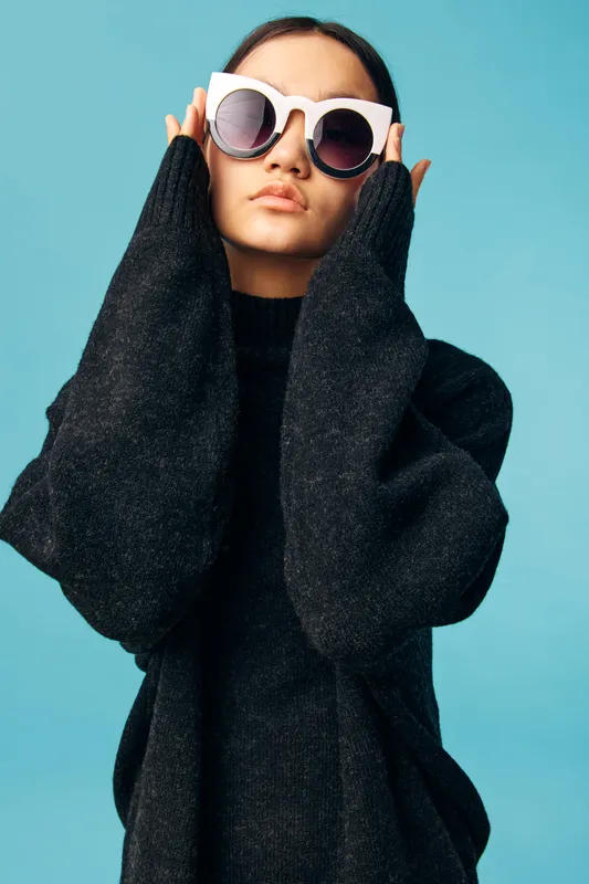 Young woman wearing oversized white sunglasses and a dark knitted sweater poses against blue background with hands near face for fashion and style concept portrait.