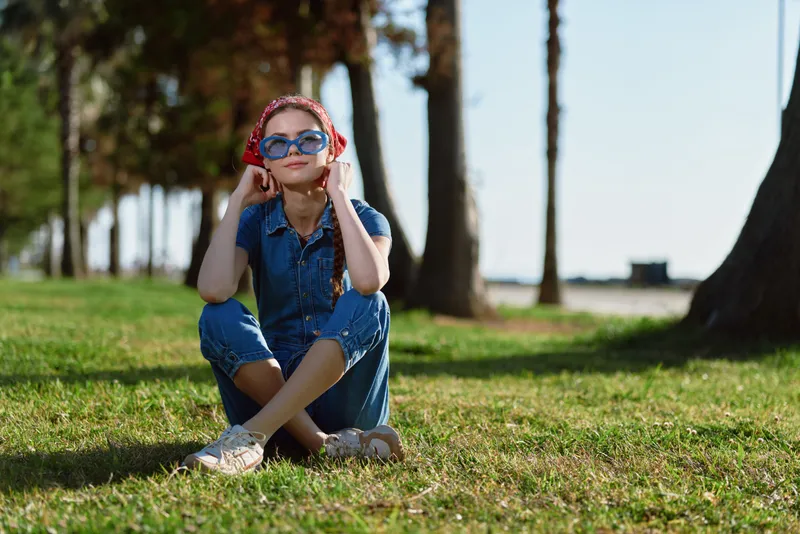 Lifestyle woman in modern boho-western denim street style with film color, wearing sunglasses and headphones, sitting relaxed outdoors on grass in sunny day.