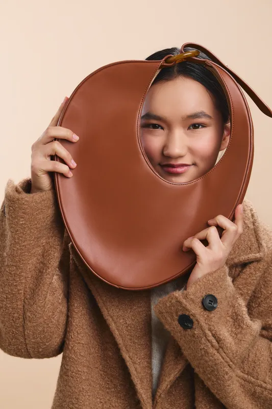 Asian woman holds a large circular bag up to her face, revealing a cheerful expression around the edges. Soft studio lighting emphasizes texture and fashion in a minimal setting.