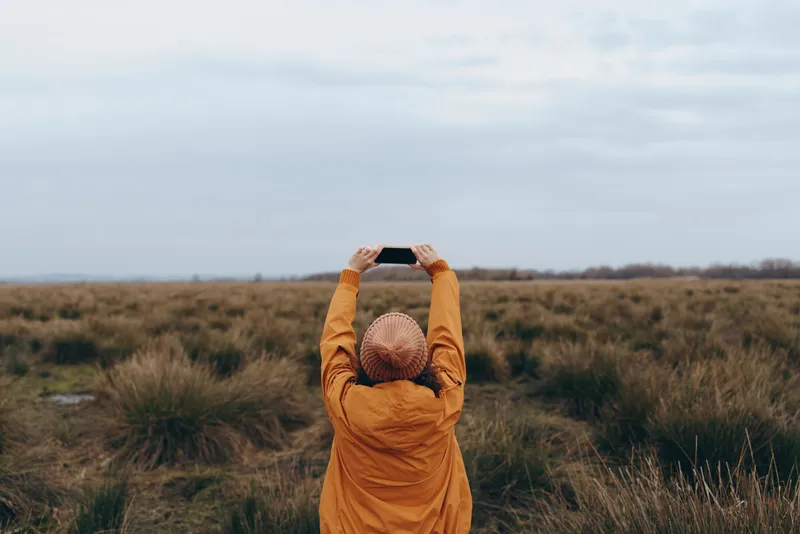 Woman lifestyle outdoors field grass sky nature calm, she bends forward holding her head in an orange coat, cloudy sky over grassy meadow, soft daylight, peaceful mood
