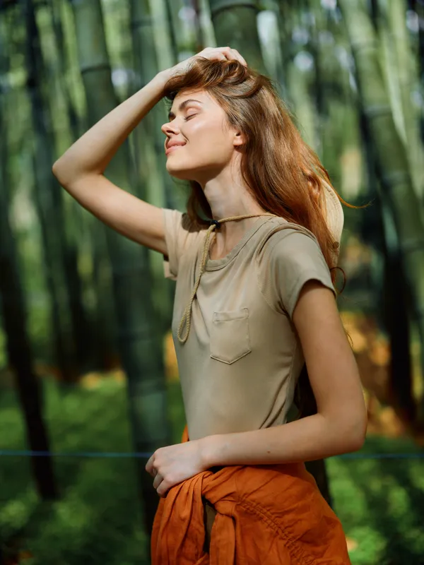 Woman portrait in forest nature, sunlight on face, relaxed pose with eyes closed and hand in hair, casual clothing, warm color tones, serene outdoor lifestyle and peaceful mood.