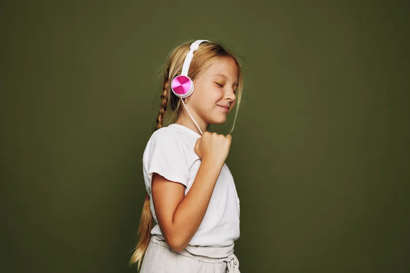 Happy girl with long hair wearing pink headphones and white t-shirt enjoys music with closed eyes and raised fist on olive background. Concept of joy and music appreciation.
