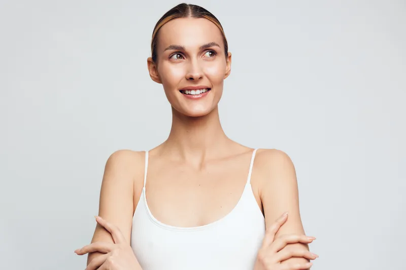 A cheerful woman poses in a studio wearing a white top, presenting a confident smile. Close up portrait with natural light, relaxed mood, and approachable beauty.