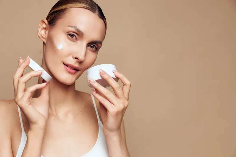 Young woman with smooth skin applying white face cream on cheek, holding open jar and tube. Neutral beige background. Concept of skincare, beauty, self-care, and daily routine.