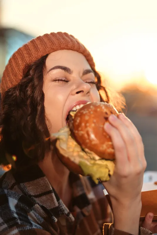 Woman biting burger in candid sunset portrait, an authentic moment with golden hour glow, mindful living and emotional storytelling of indulgence, urban comfort and enjoyment.