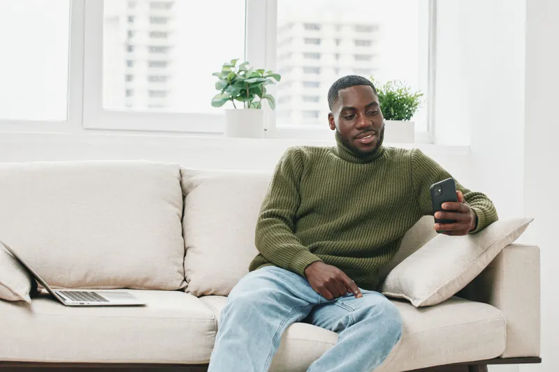 Smiling Black man relaxing on a sofa, using a smartphone, wearing a green sweater and jeans in a bright living room with plants and a laptop on the side