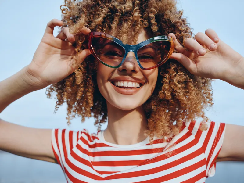 A joyful young woman with curly hair wears oversized sunglasses and a red and white striped shirt, smiling broadly against a bright blue sky on a sunny day.
