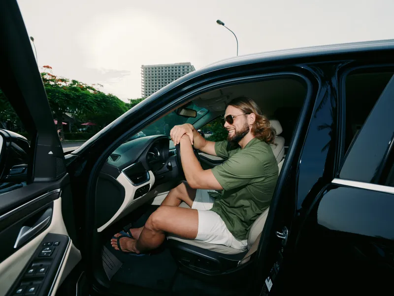 Relaxed man sits in a modern car with the door open, wearing sunglasses and a green shirt, capturing a casual summer vibe and a confident, content expression.