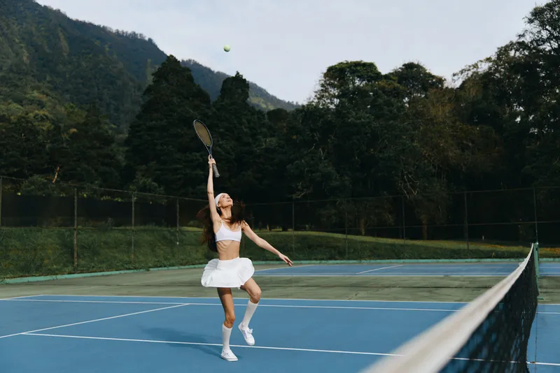 Young female tennis player in a white athletic outfit, joyfully serving on a blue court surrounded by lush greenery, capturing the spirit of athleticism and outdoor sports