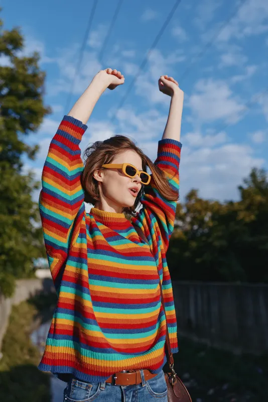 Young woman in a rainbow sweater and yellow sunglasses with arms raised outdoors, joyful expression and casual jeans outfit, crossbody bag and cozy striped knit for lifestyle fashion portrait.