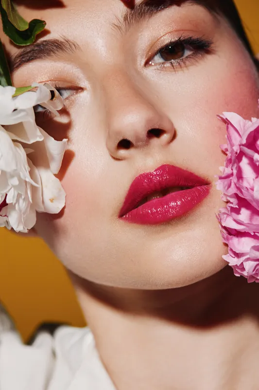 Close-up of a young woman with vibrant fuchsia lips and flawless skin, holding white and pink flowers near her face on a warm orange background. Beauty, fashion, creative concept.