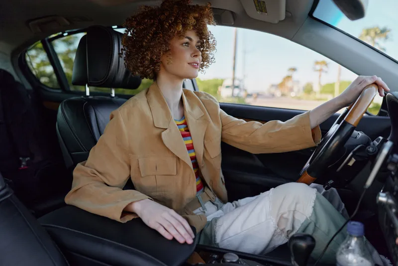 Smiling woman with curly hair driving a car wearing a tan jacket. Natural light fills the cabin, creating a warm, relaxed mood. Focus on lifestyle, fashion, and self expression during everyday travel.