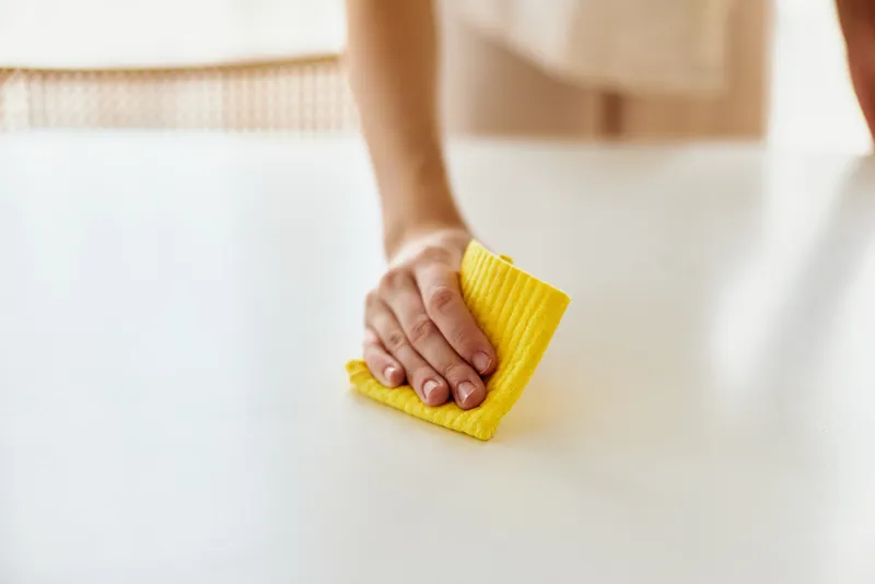 Cleaning hand using a yellow cloth to wipe a white table surface in home interior, closeup of person performing household chores and tidy maintenance for sanitation and care.