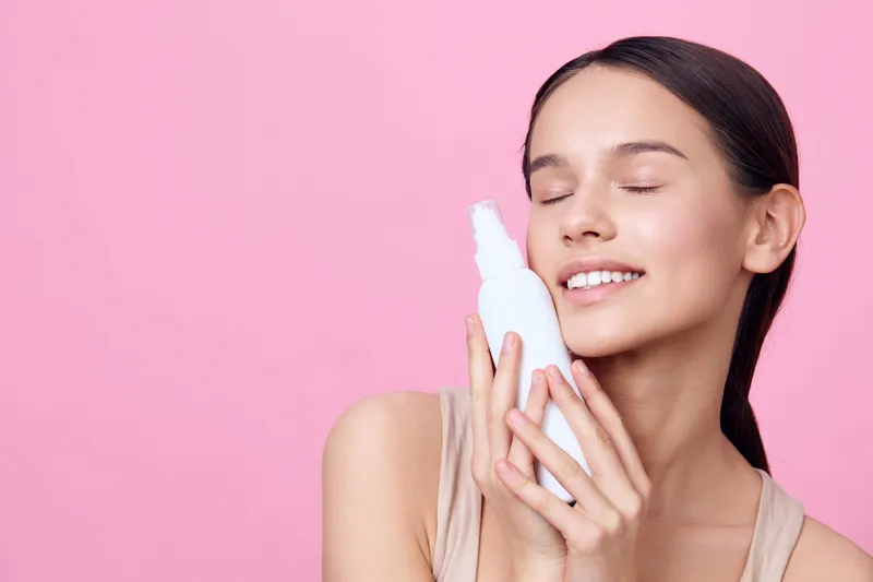 Young woman smiling with skincare product, closed eyes, pink background, expressing happiness and confidence, emphasizing natural beauty and skincare routine