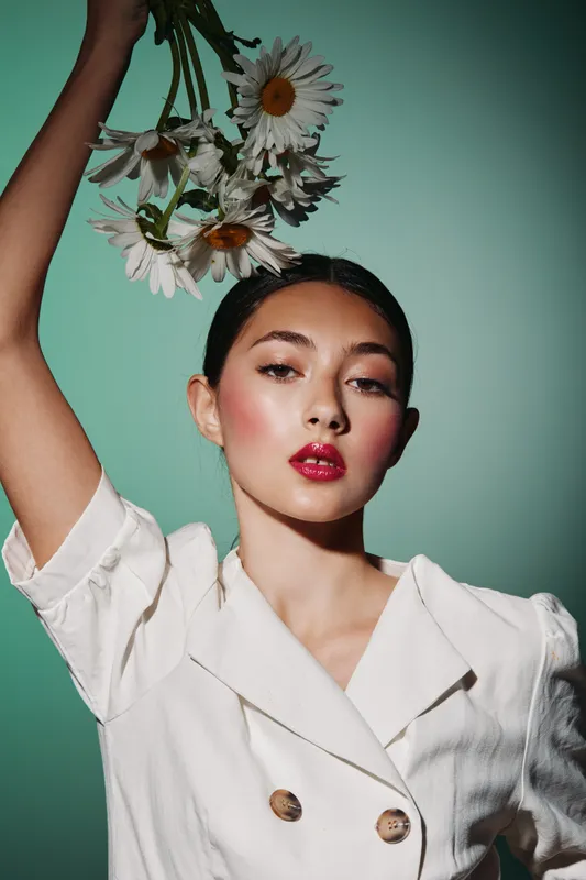 Portrait of creative woman with red lips holding fresh white daisies above her head. She wears white double-breasted clothing, posing against light green background.