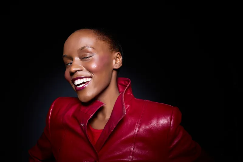 Confident black woman with dark skin wearing a red leather jacket smiles brightly against black background. Portrait of stylish African ethnicity female model with joyful expression and glamour.