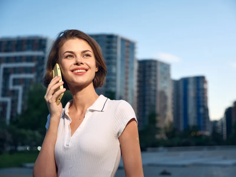 A cheerful woman is talking on a smartphone in an outdoor urban setting. She smiles warmly, projecting confidence and a modern, dynamic lifestyle in a bright city scene.