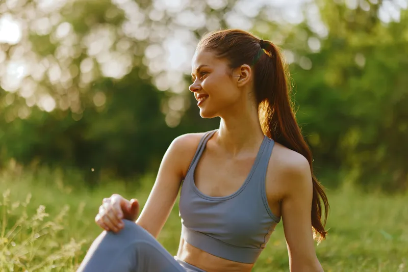 Young athletic woman in gray sportswear enjoying nature with a joyful expression in a sunlit green setting, promoting healthy lifestyle and fitness