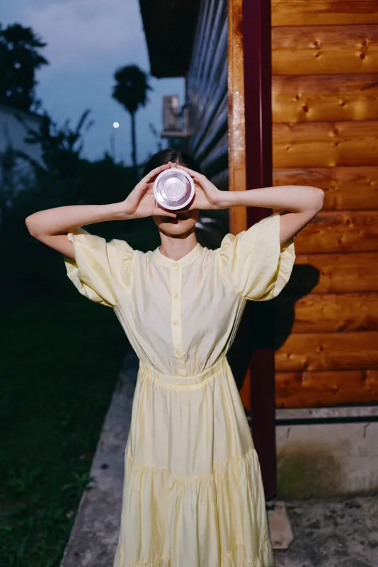 Portrait of a woman in a pale yellow dress outdoors at dusk, holding a cup up to her eyes. The scene captures a playful, mysterious moment against a wooden exterior and evening light.