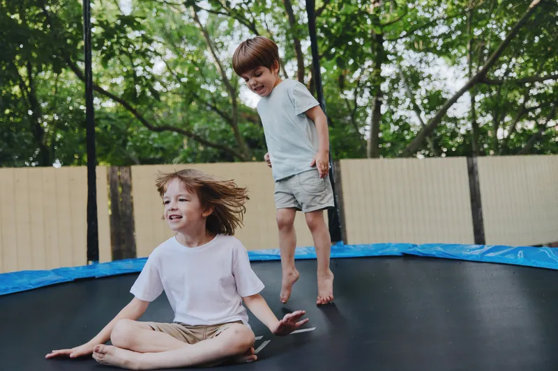 Happy children jumping on a trampoline, enjoying a sunny day outdoors surrounded by green trees. They express joy and excitement in a playful atmosphere.