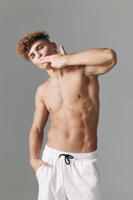 Young muscular man with curly hair posing shirtless wearing white shorts with drawstring and hand in pocket. Studio background and expression with relaxed mood and fit body.