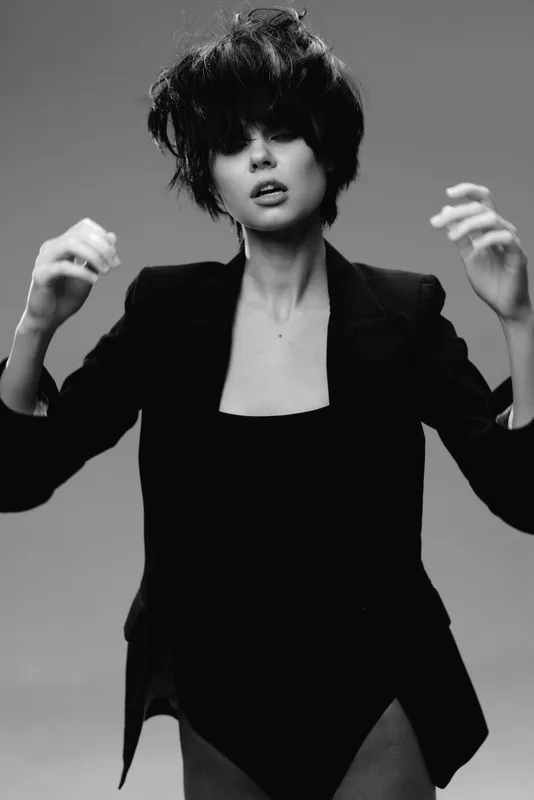 Black and white portrait of a stylish woman with short hair and expressive pose in dark clothing against plain background.
