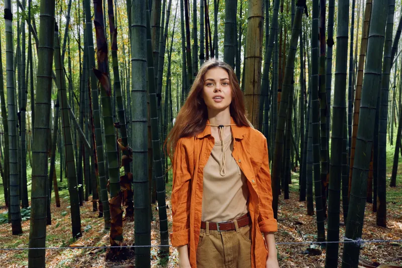 woman bamboo forest portrait standing nature fashion outdoors, young model in orange jacket and linen blouse poses among tall green stalks, serene expression and natural light