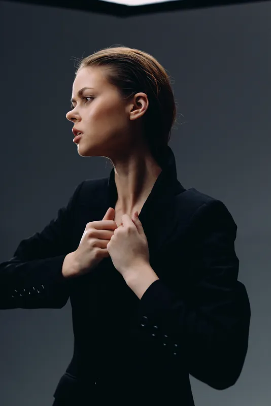 Stylish woman adjusting black jacket with serious expression in dark studio setting