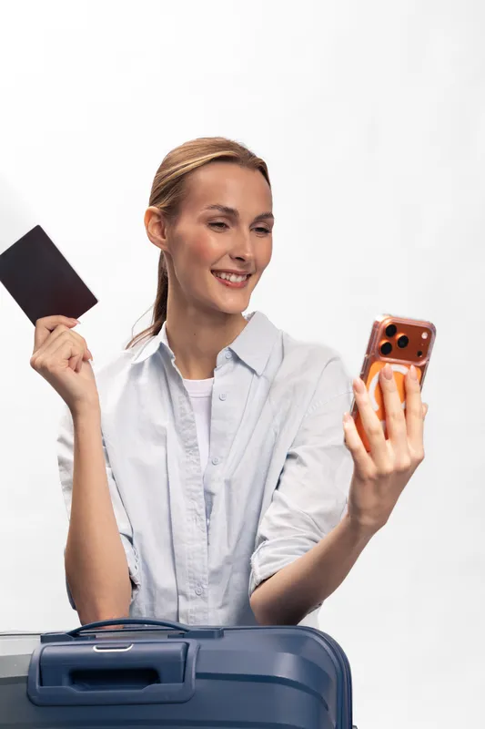 Smiling woman holds passport and smartphone while standing behind suitcase. Concept of travel preparation, vacation planning, and modern tourism with happy female traveler in casual clothes.