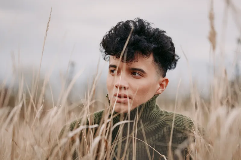 Candid portrait of a young person amid tall dry grass, contemplative gaze and soft natural light conveying authenticity and genuine emotion, close framed, neutral tones and textured surroundings.