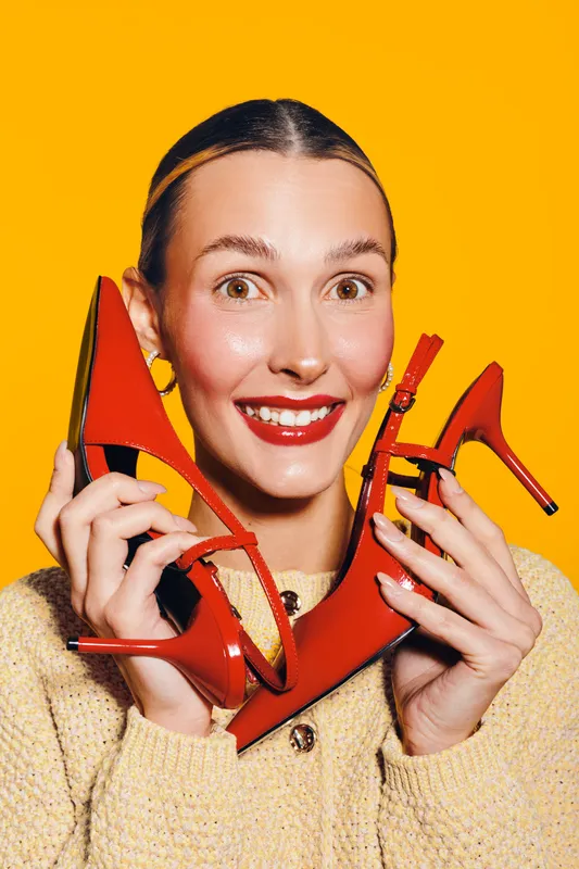 Happy woman with red high heels smiles excitedly on yellow background. Concept of fashion, style, shopping, and joyful emotions with bright colors and trendy footwear.