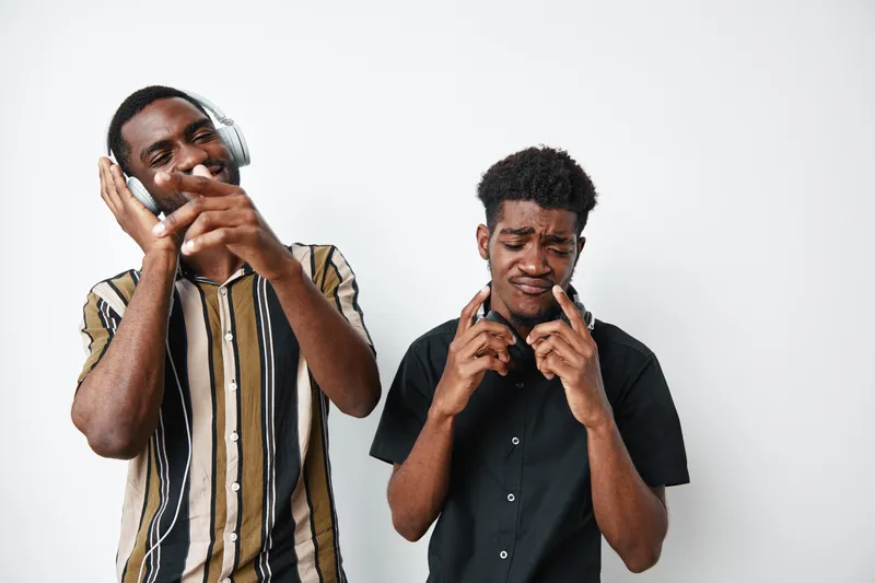 Two young black men with dark skin enjoying music together, one wearing headphones and striped shirt, the other in black shirt, expressing emotions and having fun on white background.