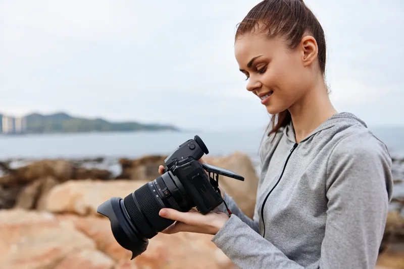 Young woman smiling while reviewing photos on a camera by the seaside, dressed in casual sportswear, showcasing a joyful and relaxed atmosphere