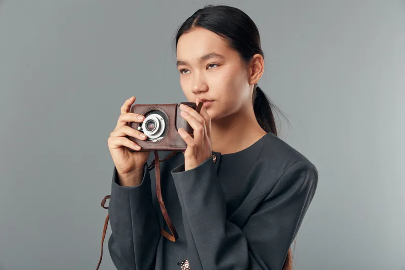 Woman with long hair holding vintage camera for photography and retro style with focused expression on face. Studio portrait with minimalistic grey background and casual dark clothing.