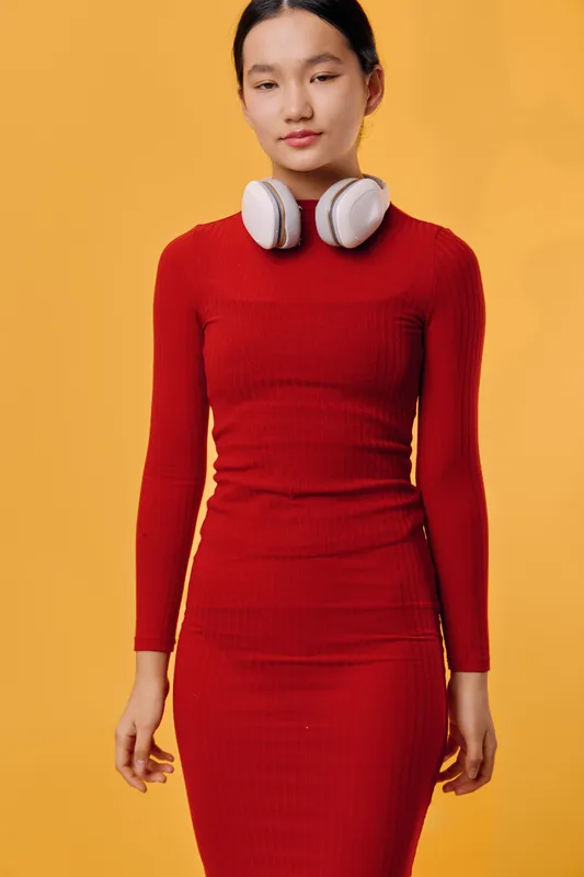 Young woman with headphones around neck wearing red dress standing on yellow background with eyes closed, calm expression and relaxed posture in studio portrait.