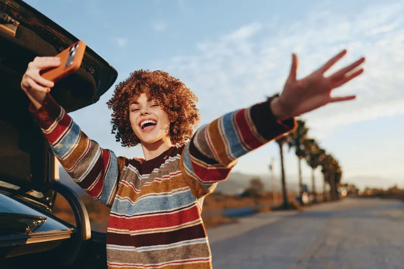 Woman lifestyle, curly hair girl near car at sunset, holding smartphone and smiling widely, arms open on road, striped sweater, joyful outdoor moment, golden sky background