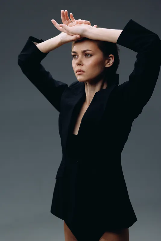 serious woman in black blazer posing with arms raised against gray background, fashion, portrait, elegant