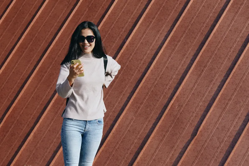 Woman in casual attire posing outdoors with sunglasses and smartphone against a wooden wall, street fashion and confident lifestyle shot for modern urban style, sunny mood and everyday chic.