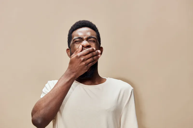 A young African man yawning with a neutral expression, wearing a light colored t shirt against a soft beige background, reflecting fatigue and relaxation