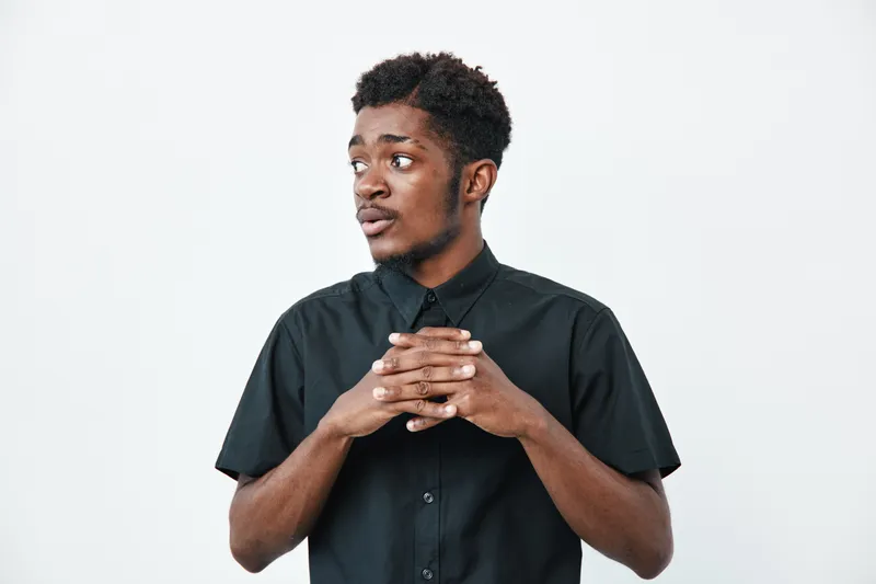 Young black man with dark skin wearing black shirt standing with hands clasped looking sideways on white background. Portrait of thoughtful African American male in casual clothing.