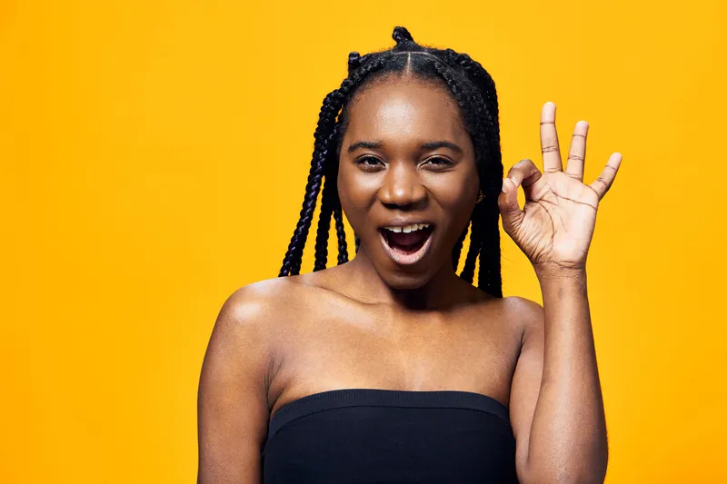 Young Black woman with braided hair showing an OK gesture against a vibrant yellow background, exuding confidence and joy, perfect for positive messaging