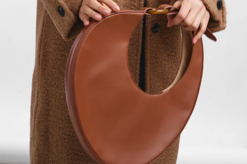 Close up of a brown circular handbag held by a gloved hand beside a textured coat, showcasing chic fashion and everyday style in a clean studio setting.