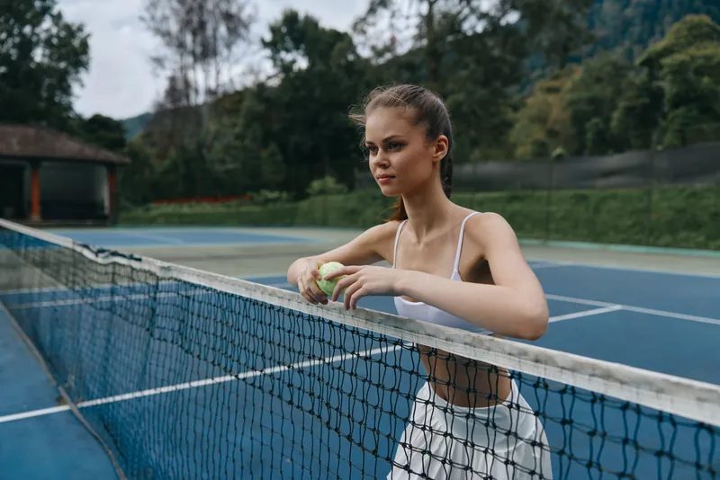 Young female tennis player in white sportswear gazing thoughtfully over the net on a blue court, embodying concentration and athleticism in a natural setting