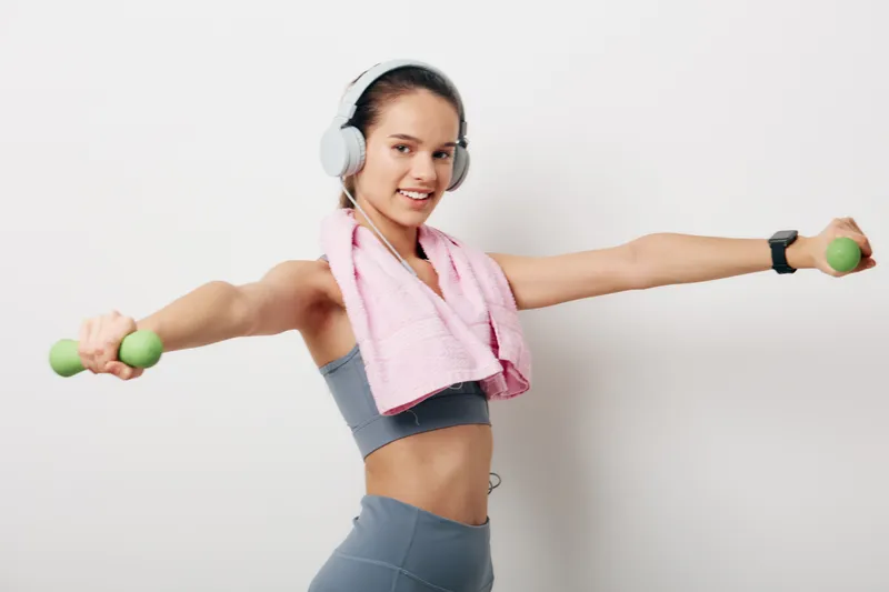 Young woman fitness portrait exercising with small green dumbbells, wearing headphones and a pink towel around her neck, grey activewear and smartwatch, smiling against a light studio background.