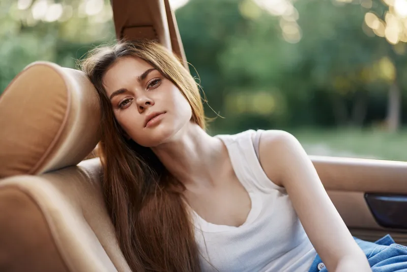 Young woman with long hair relaxing in car during sunny day, showcasing a serene expression and casual clothing in a natural outdoor setting.