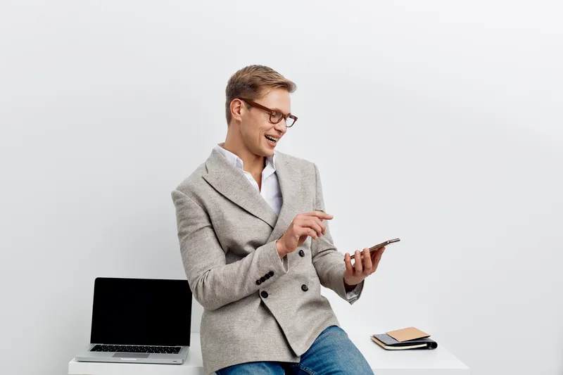 Smiling man using smartphone sitting at modern workplace with laptop and notebooks in bright office environment. Confident professional male engaging with mobile device, casual business attire, positi