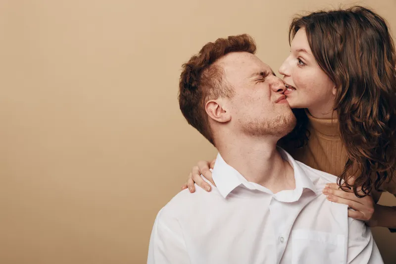 Young couple shares a playful moment as woman kisses man on cheek. Both show affection and joy in a warm, neutral background. Concept of love, intimacy, and happiness.