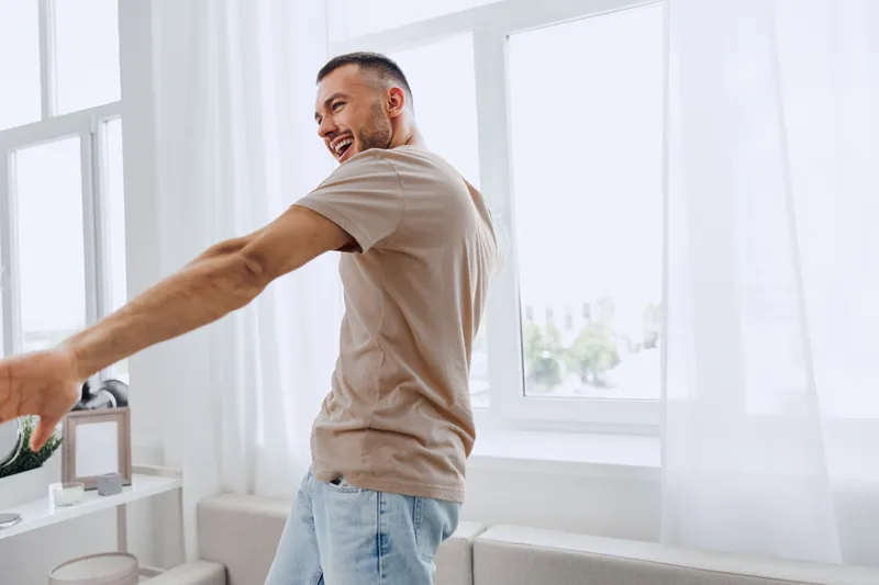 Smiling man joyfully dancing at home, wearing a casual beige t-shirt and light jeans. Bright, airy atmosphere with soft natural light creates a cheerful vibe.